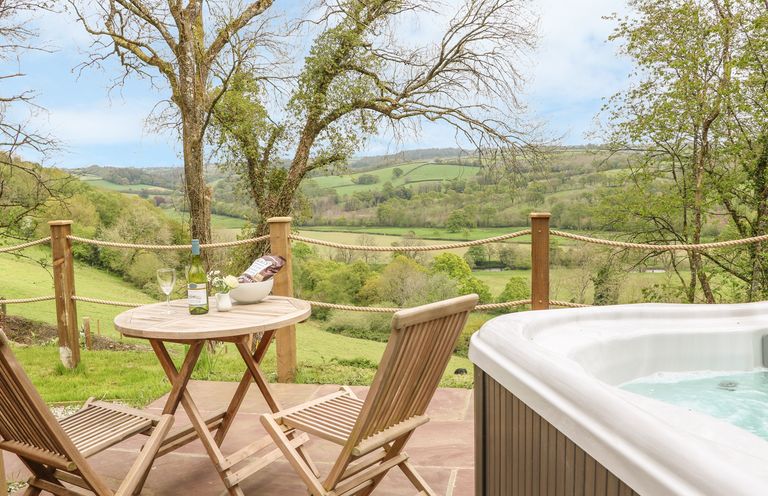 An outdoor patio with two wooden chairs and a table with a bottle of wine and glass next to a hot tub overlooking green hills at Little Silver Owl in High Bickington
