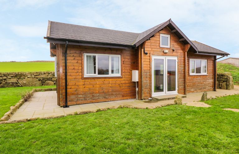 A wooden cabin with white framed windows and doors on a stone patio surrounded by grass at Borah Lodge in Mousehole