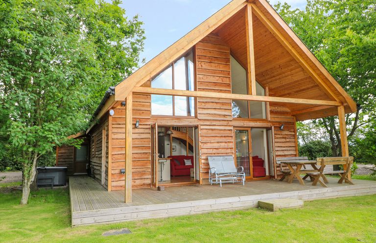 A wooden cabin with a deck, patio furniture, and trees surrounding it at Dorado Lodge in Thorpe-on-the-Hill