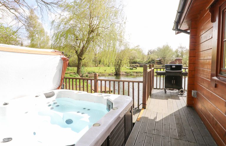 An outdoor wooden deck with a hot tub and a barbecue grill next to a pond surrounded by trees at Heron Lodge in Badwell Ash