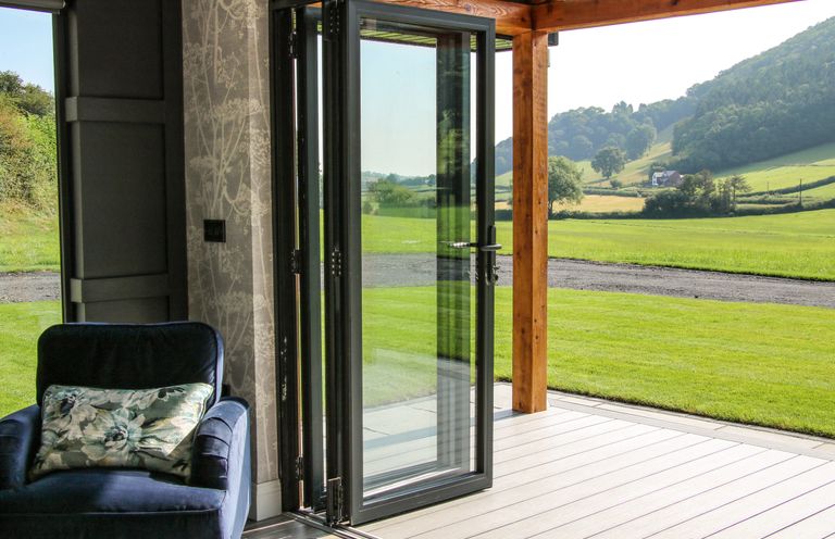 An armchair with a floral cushion next to a folding glass door opening to a patio and green fields at Meadow View in Meifod Powys