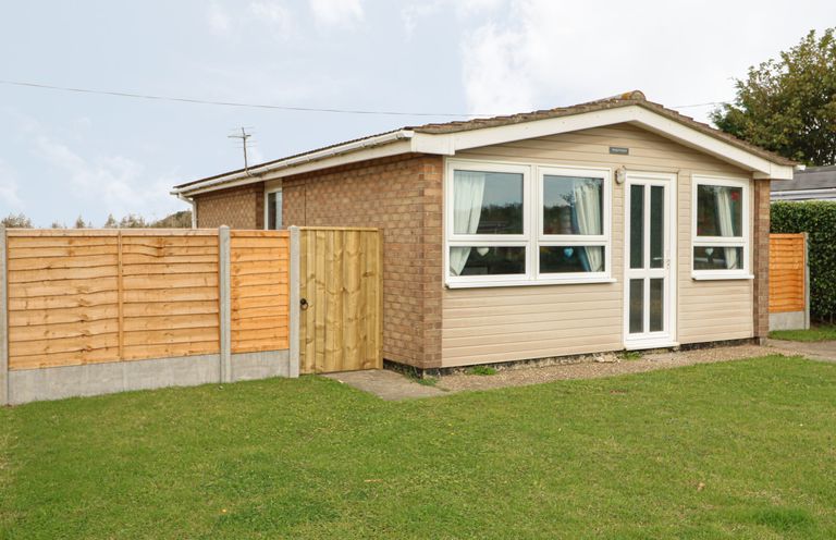 A small single-story house with a front door and windows with curtains surrounded by a wooden fence and green lawn at Moggs Retreat in Chapel St Leonards