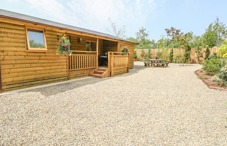 A wooden cabin with a small porch, hanging flower baskets, a gravel yard, wooden picnic table, and surrounding plants at Willow Lodge in St Florence