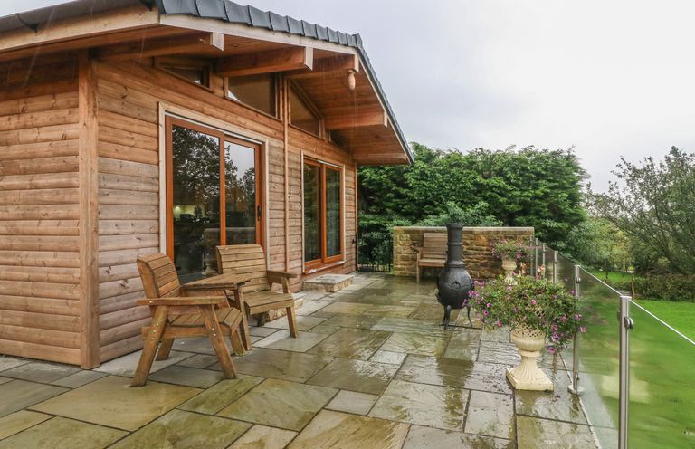 A wooden cabin with glass doors a stone patio wooden chairs a chiminea glass railing and flower pots at Nicky Nook Lodge in Scorton