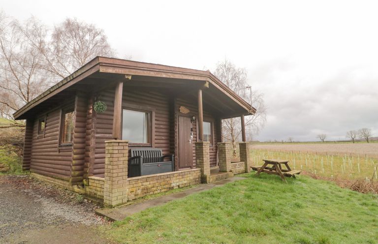 A small log cabin with a covered porch and bench next to a grassy area with picnic table at Glanton in Longframlington