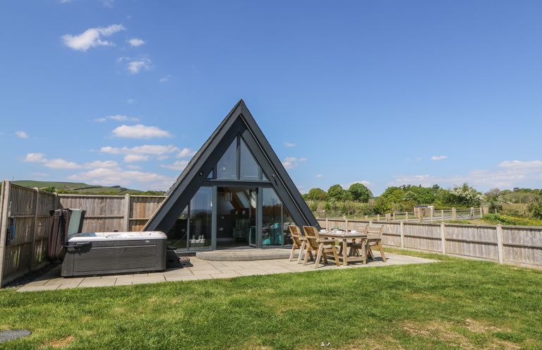 An outdoor patio with a hot tub and wooden table with chairs next to an a frame building at Elan Valley Welsh - The Sheepfold in Llanyre near Llandrindod Wells