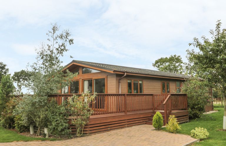 A wooden lodge with a deck and railings surrounded by trees and bushes at Bulmer Farm Lodge in Malton