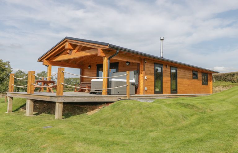 An exterior view of a wooden log cabin with a covered porch and picnic table on a grassy lawn at Bryn Eiddon Log Cabin in Machynlleth