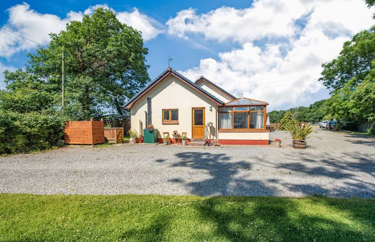A white house with a wooden door and a glass conservatory next to a gravel driveway with plants and trees in the background at Covert Farm Cwtch in Jeffreyston