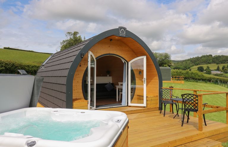 A wooden pod with open doors on a deck with a hot tub and two black chairs in a green field at Seren in Llandrindod Wells