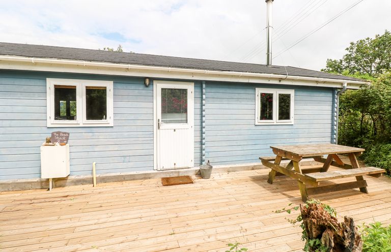 An exterior of a blue wooden cabin with white door and windows and a wooden picnic table on a deck at The Beach Shack in Llansteffan