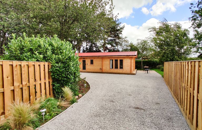 A wooden cabin at the end of a gravel driveway with wooden fences and trees on both sides at Gardener's Lodge in Huby