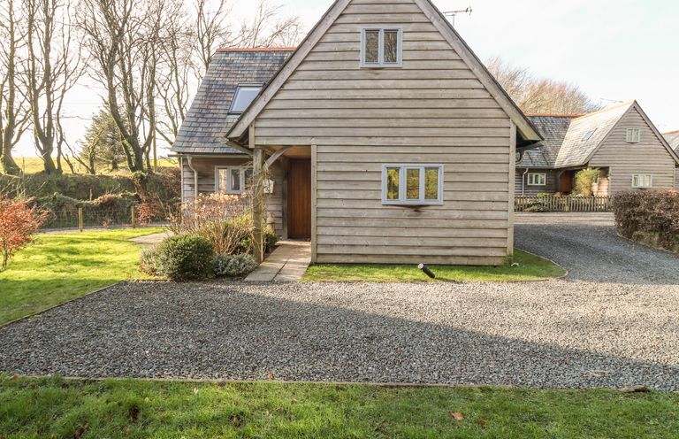 A wooden house with a gravel driveway and grass lawn surrounded by trees at Honeypot Lodge in Camelford