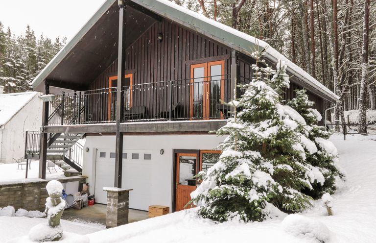 A snowy house with a balcony and garage doors surrounded by snow-covered trees at Torbreck Chalet in Kincraig