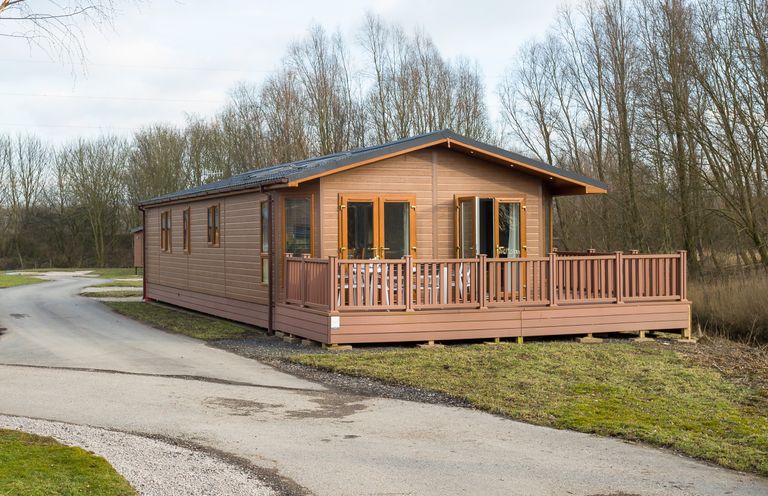 A wooden lodge with a railing around a porch on a grassy plot near a road at Waternook Lodge in South Lakeland Leisure Village