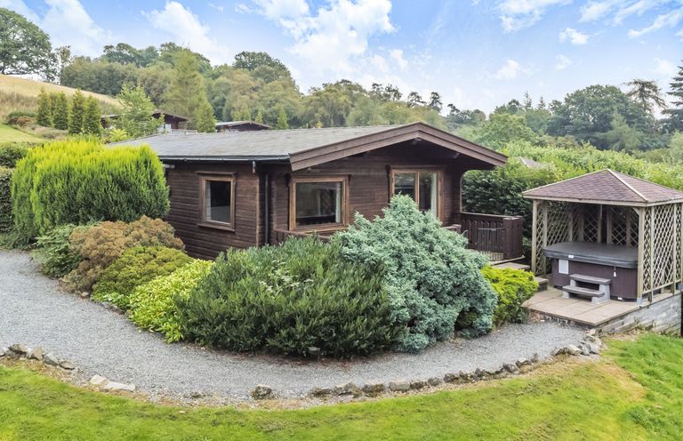 A small wooden cabin surrounded by bushes and trees with a gazebo containing a hot tub nearby at Hillside View in Rhayader