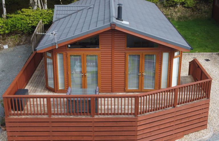 A wooden lodge with a gray roof surrounded by a wooden deck and fence in a green area at Lodge 11 in Tavistock