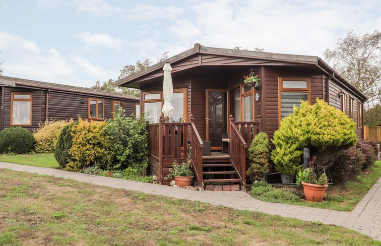 A wooden cabin with a porch and steps surrounded by bushes and a paved walkway at 4 Country View Park in Graveney near Seasalter