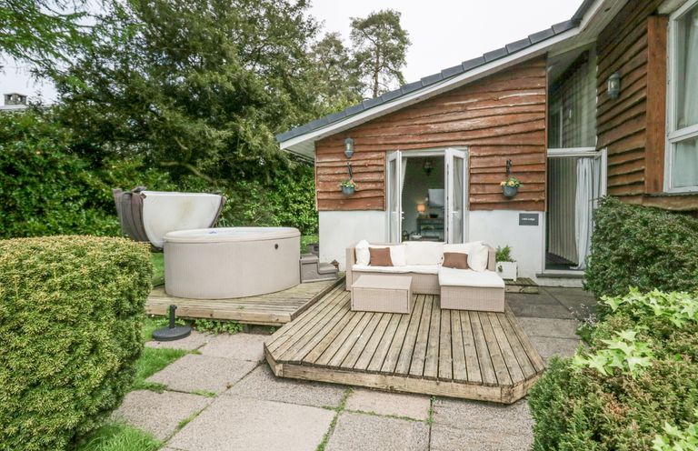 An outdoor patio with a wooden deck sofa hot tub and surrounding bushes at Lake Lodge in Bowness-On-Windermere
