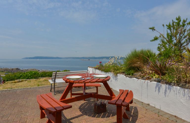 An outdoor patio with a round wooden picnic table set with plates and glasses overlooking the sea at SoundView in Down Thomas
