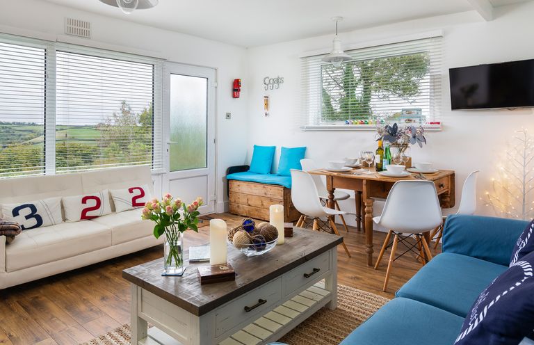 A living room with a white sofa and blue cushions a wooden coffee table and a dining table with white chairs and tableware at The Hut in Dartmouth
