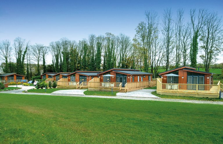 A row of wooden lodges with decks and gravel paths surrounded by trees and grass at Otter Lodge in Gargrave