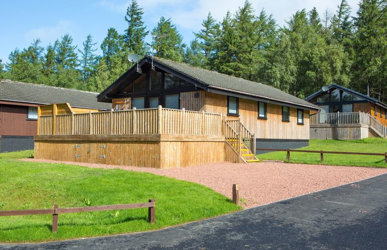 A wooden cabin with a fenced porch and gravel driveway surrounded by grass and trees at Pine Tree in Otterburn