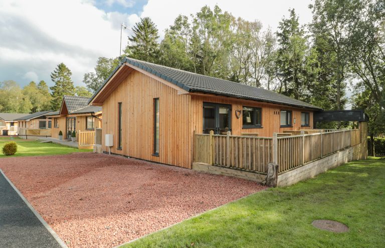 A single-story wooden house with a fenced porch and gravel driveway surrounded by grass and trees at Barn Owl in Otterburn