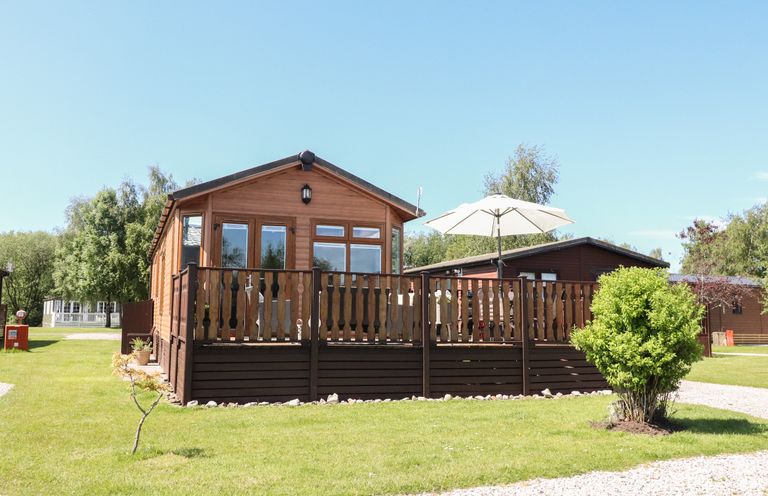 A wooden cabin with a deck and umbrella in a grassy area at Doormouse Lodge in South Lakeland Leisure Village