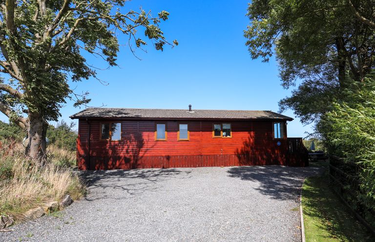 A red wooden cabin with multiple windows surrounded by trees and a gravel driveway at Ffrwd Lodge in Rhosneigr