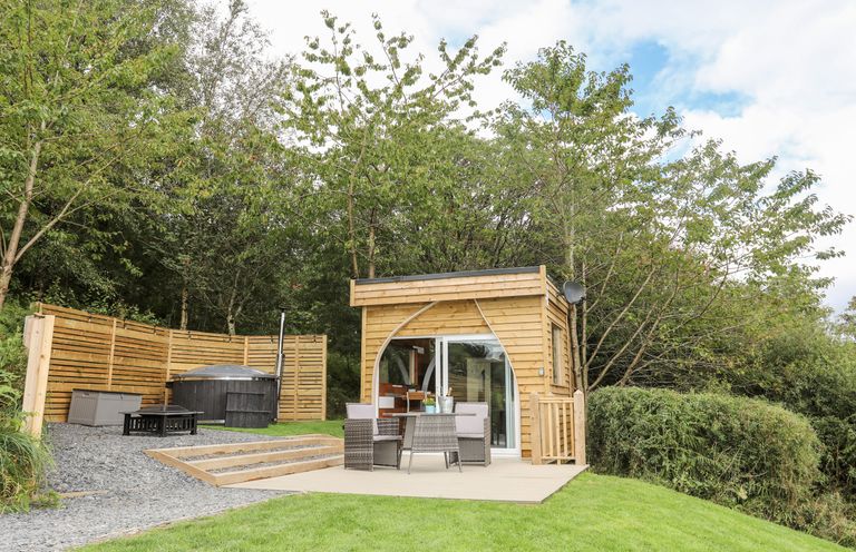 A small wooden cabin with outdoor dining furniture and a hot tub surrounded by trees at Woodland Pod Maerdy near Corwen