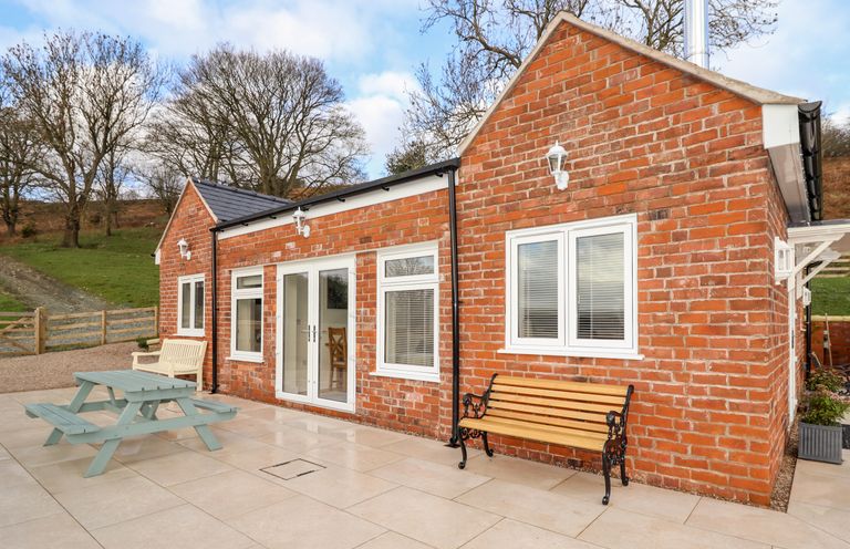 An outdoor patio area with a brick building a wooden bench a picnic table and wooden chairs at Border View Lodge in Llandrinio