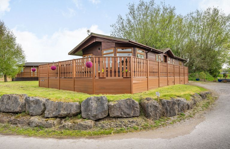 A wooden lodge with a fenced porch on a grassy area with large rocks and trees at Lakeview Lodge in South Lakeland Leisure Village