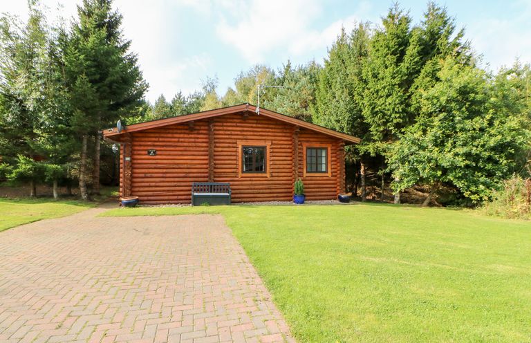 A log cabin with two windows and a bench in front on a paved driveway surrounded by grass and trees at Jalan End in Felton