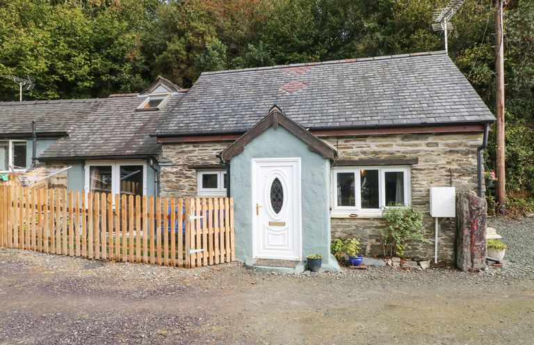 A stone cottage with a white door and wooden fence surrounded by trees at Pendre Cottage in Cenarth