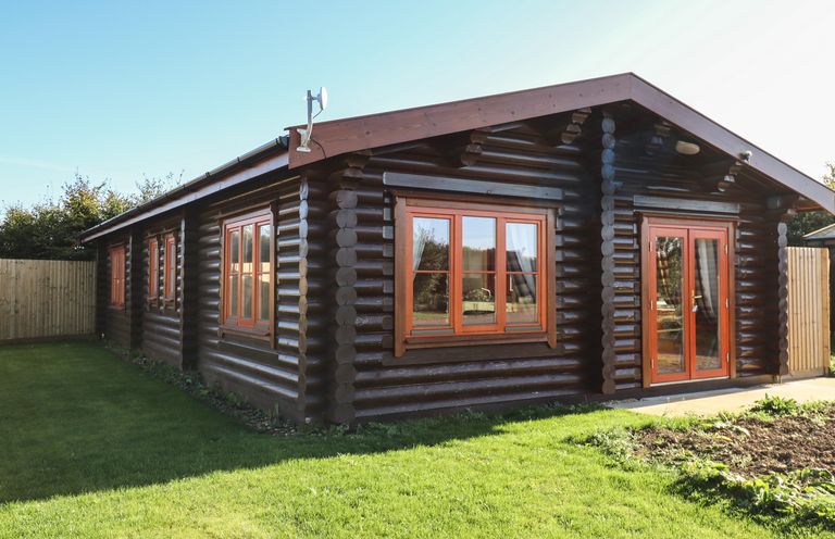 A log cabin with multiple windows and a glass door surrounded by grass and fenced yard at Rowan Lodge in Greetham Rutland