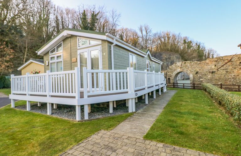 A wooden cabin with white railings on a raised platform near a stone archway and trees at The Hideaway - Heritage Park in Stepaside near Kilgetty