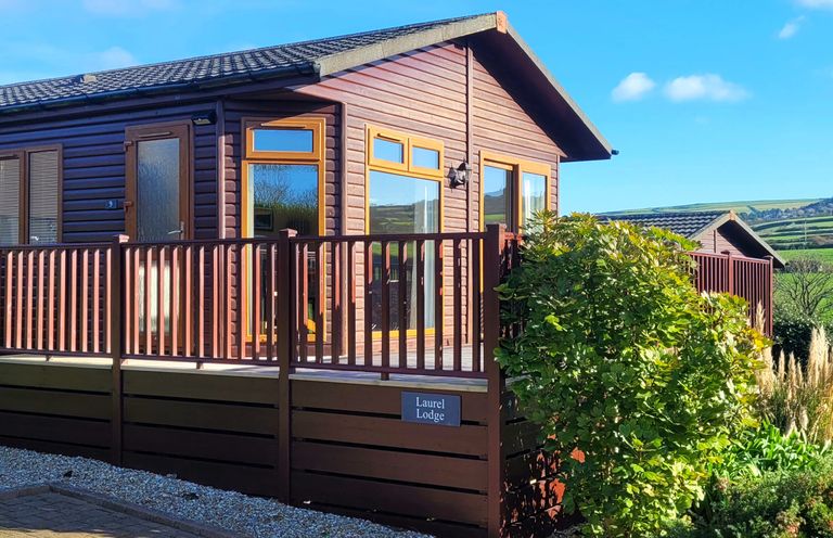 A wooden lodge with a deck and railing surrounded by bushes at Laurel Lodge Bossiney Bay Holiday Park near Tintagel