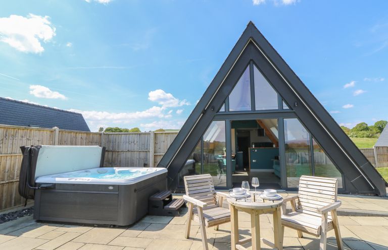 An outdoor patio with a hot tub and wooden table with chairs near a glass door of an A-frame building at Kerry Hill - The Sheepfold in Llanyre near Llandrindod Wells