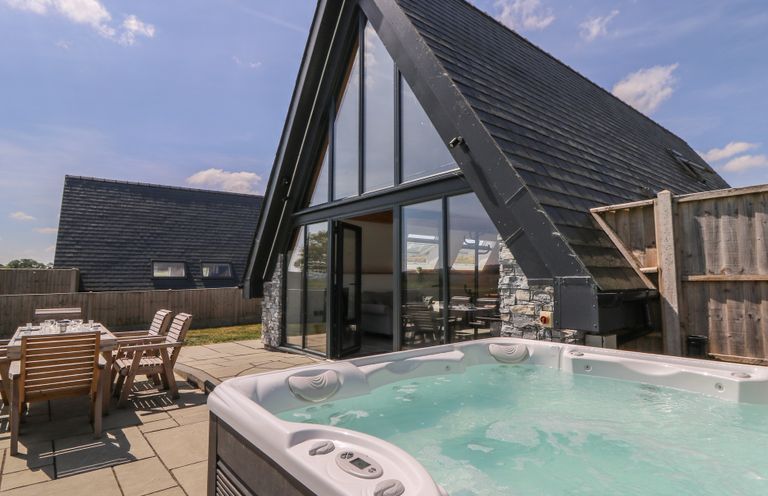 An outdoor patio with a hot tub and wooden dining table with chairs next to a house with large glass windows at Hill Radnor - The Sheepfold in Llanyre near Llandrindod Wells