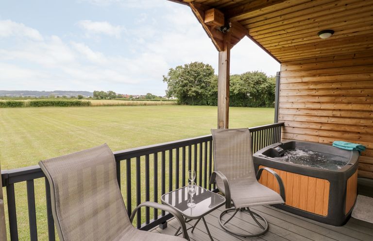 An outdoor covered deck with two chairs a small table with glasses and a hot tub overlooking a grassy field at Cotswold View 1 in Honeybourne