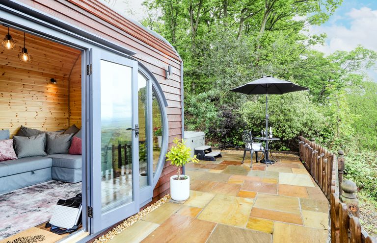 A wooden cabin with glass doors opening to a stone patio with a table umbrella and chairs surrounded by trees at Offas Dyke Escape in St Asaph