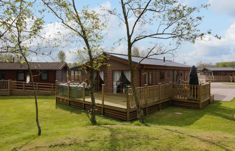 A wooden lodge with a deck and outdoor seating surrounded by grass and trees at Lakeland Dream @ South Lakeland Leisure Lodge in Warton