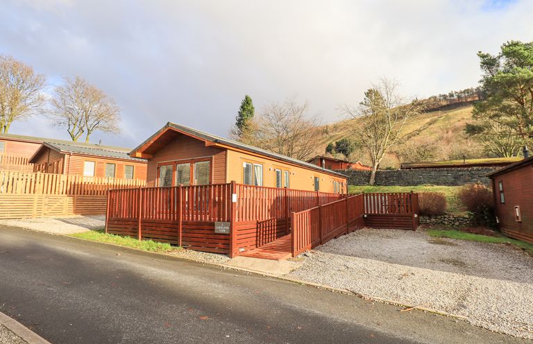A wooden lodge with a red wooden ramp and railings next to a gravel parking area with trees and hills in the background at Lakeland View Lodge Beckside 2 Limefitt Park near Windermere