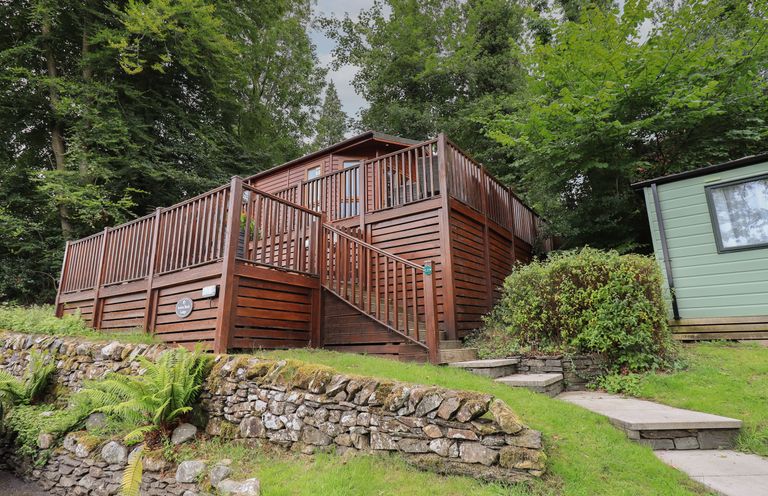 A wooden lodge with a deck and stairs surrounded by greenery and stone walls at Acorn Bank Lodge L 55 Fallbarrow Park near Bowness-On-Windermere