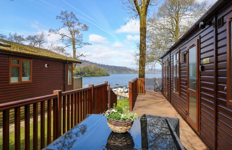 A deck with wooden railings and a table with a flower basket between two wooden lodges by a lake at Shoreside Lodge L8 Fallbarrow Park near Bowness on Windermere