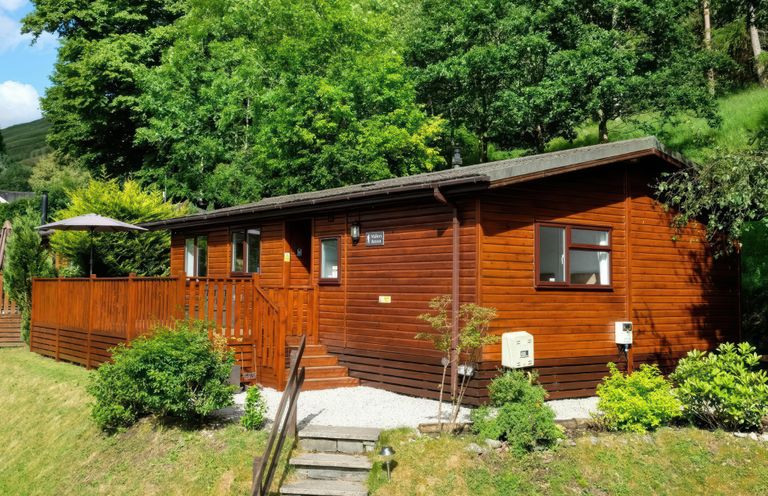 A wooden cabin with a deck and umbrella surrounded by trees and bushes at Walkers Retreat Lodge Fellside 5 Limefitt Park near Windermere