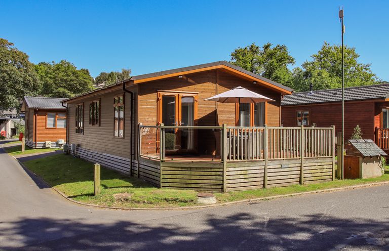A wooden cabin with a fenced deck and umbrella surrounded by trees and other cabins at Daffodils Lodge Oakside 7 Fallbarrow Park in Bowness on Windermere