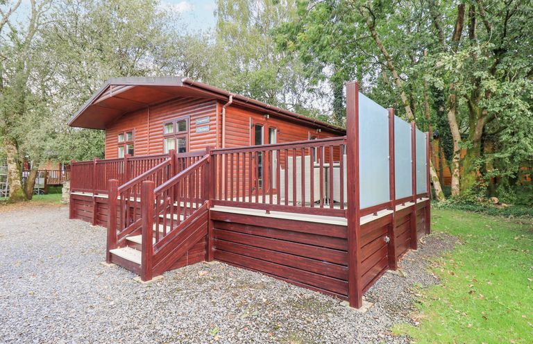 A wooden lodge with a raised deck and stairs surrounded by trees and gravel at Tanglewood Lodge Thirlmere 15 White Cross Bay near Windermere