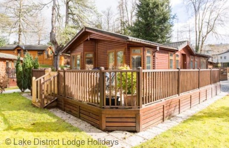A wooden lodge with a fenced deck surrounded by grass and trees at Broad Larch Lodge Glade 16 Fallbarrow Park near Bowness on Windermere